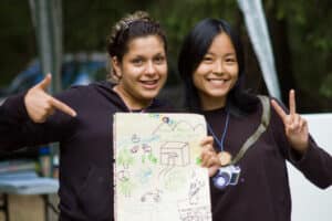 Two youth smiling at camera, holding a hand-drawn image of a community center, school, and other sustainable elements