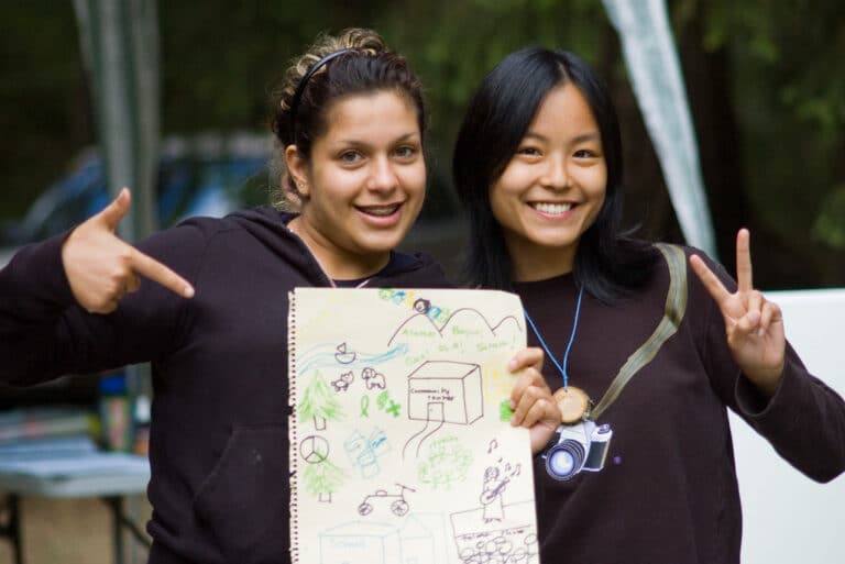 Two youth smiling at camera, holding a hand-drawn image of a community center, school, and other sustainable elements